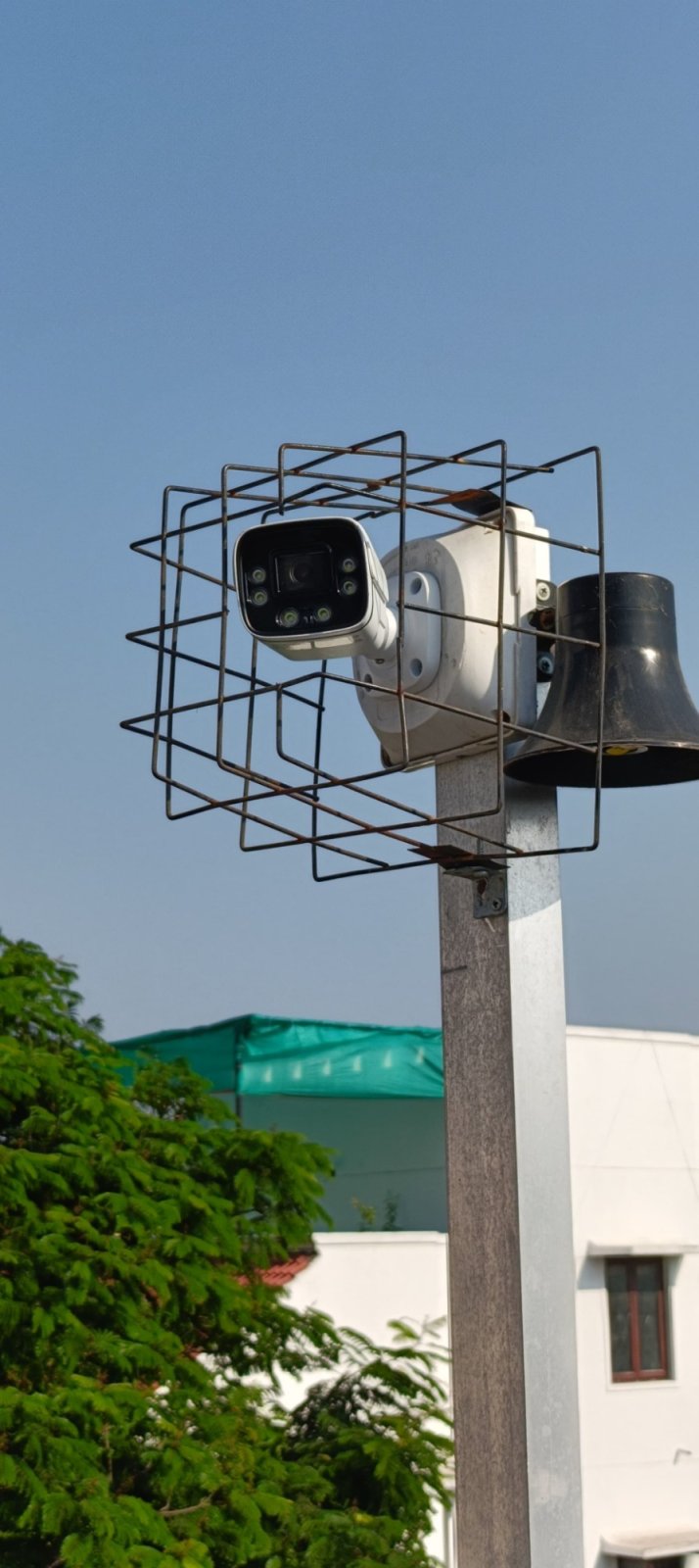 Outdoor CCTV camera inside a protective metal cage next to a siren horn speaker on a pole